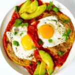 An overhead look at a plate of Huevos Rancheros. The sauce is bright red, the eggs are sunny-side-up and there are avocado slices on the plate as well.