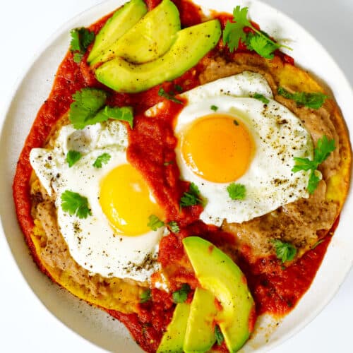 An overhead look at a plate of Huevos Rancheros. The sauce is bright red, the eggs are sunny-side-up and there are avocado slices on the plate as well.