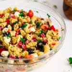 A big, glass mixing bowl full of Italian Pasta Salad sits on a white work surface. It is vibrant and full of color.