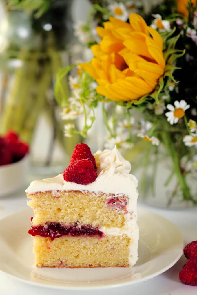 A piece of Lemon Raspberry Cake sits on a white plate with a bouquet of sunflowers and greenery behind it.