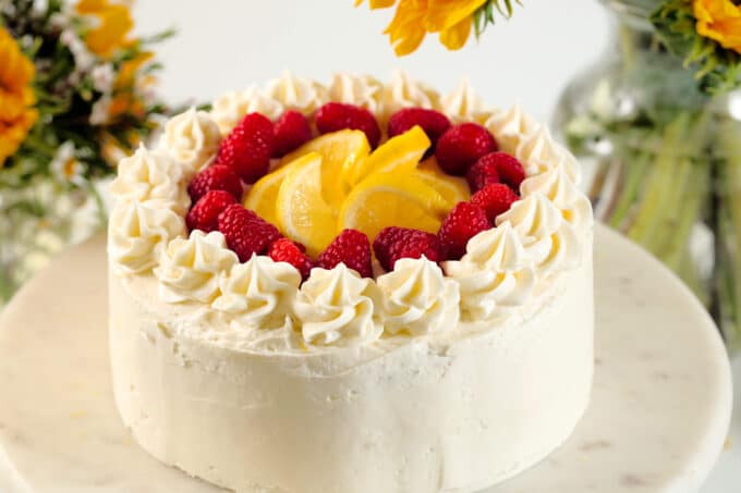 A whole Lemon Raspberry Cake sits on a cake stand. It has frosting piped around the top with lemon wheels and fresh raspberries in the middle.