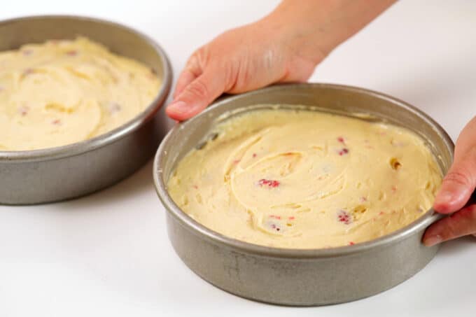 Hands holding a cake pan filled with Lemon Raspberry Cake batter. The other pan of batter is sitting on the counter beside it.