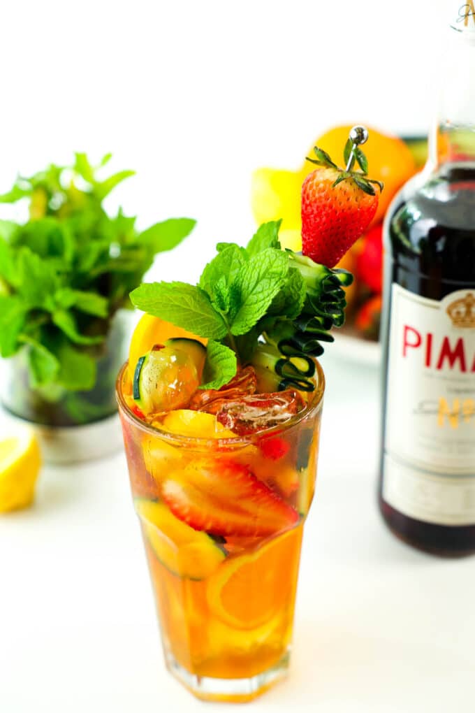 A Pimm's Cup cocktail sits on a counter with a bottle of Pimm's and other ingredients sitting behind it. The glass has fruit mixed in and a garnish of mint, a strawberry, and a cucumber ribbon on a skewer. 