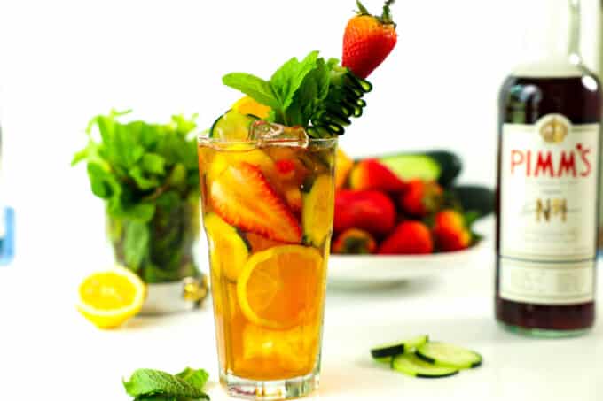 A Pimm's Cup cocktail sits on a counter with a bottle of Pimm's and other ingredients sitting behind it. The glass has fruit mixed in and a garnish of mint, a strawberry, and a cucumber ribbon on a skewer. 