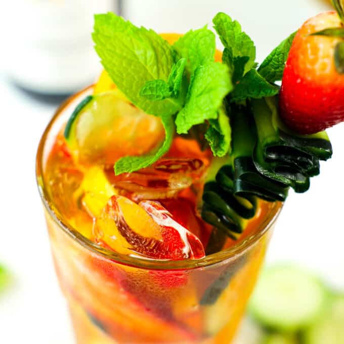 A close up of the top of a Pimm's Cup with a garnish of mint leaves, and a cucumber ribbon on a skewer with a whole strawberry on top.