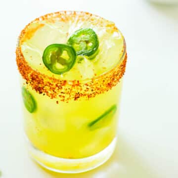 A close up of a Spicy Margarita in a rocks glass, with a Tajin rim and slices of jalapeño on top of the ice and throughout the glass.