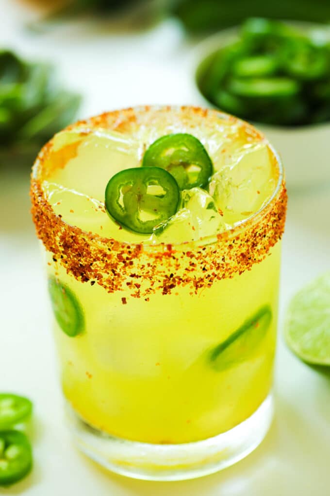 A close up of a Spicy Margarita in a rocks glass, with a Tajin rim and slices of jalapeño on top of the ice and throughout the glass. There are sliced jalapeños in bowls and on the counter around it.
