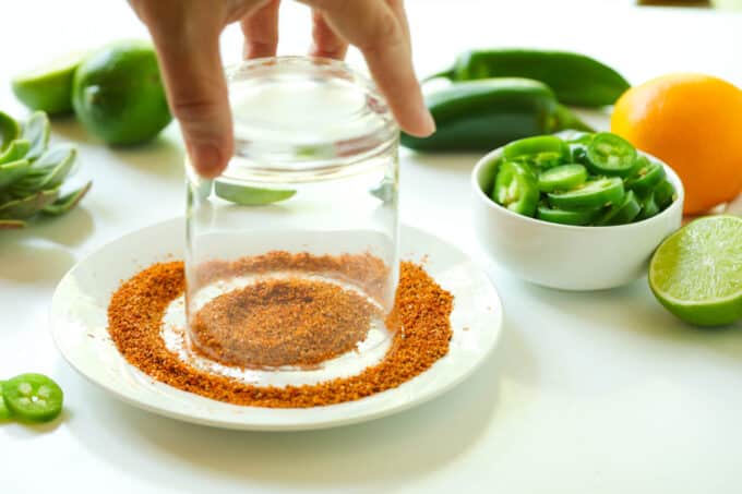 A rocks glass upside down on a plate of Tajin. A hand is holding the bottom of the glass to swirl it around in the Tajin.