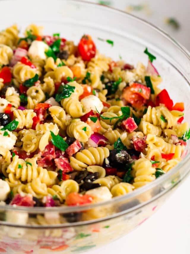 A big, glass mixing bowl full of Italian Pasta Salad sits on a white work surface. It is vibrant and full of color.