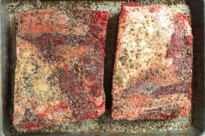 Two racks of raw Beef Ribs laying side by side with salt and pepper sprinkled over them.