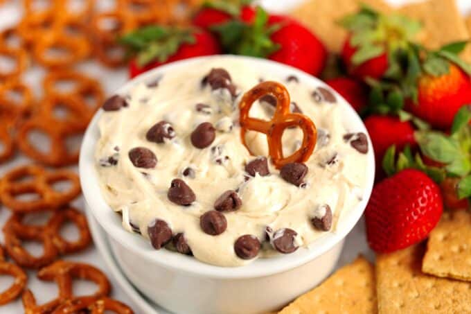 A white bowl full of Cookie Dough Dip surrounded by pretzels, strawberries, and graham crackers, with one pretzel stuck in the dip.