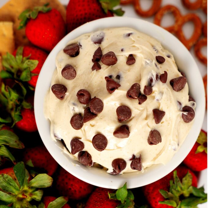 An overhead view of a bowl of Cookie Dough Dip. It is a light, off-white color and has chocolate chips mixed in. There are strawberries and pretzels sitting around it.