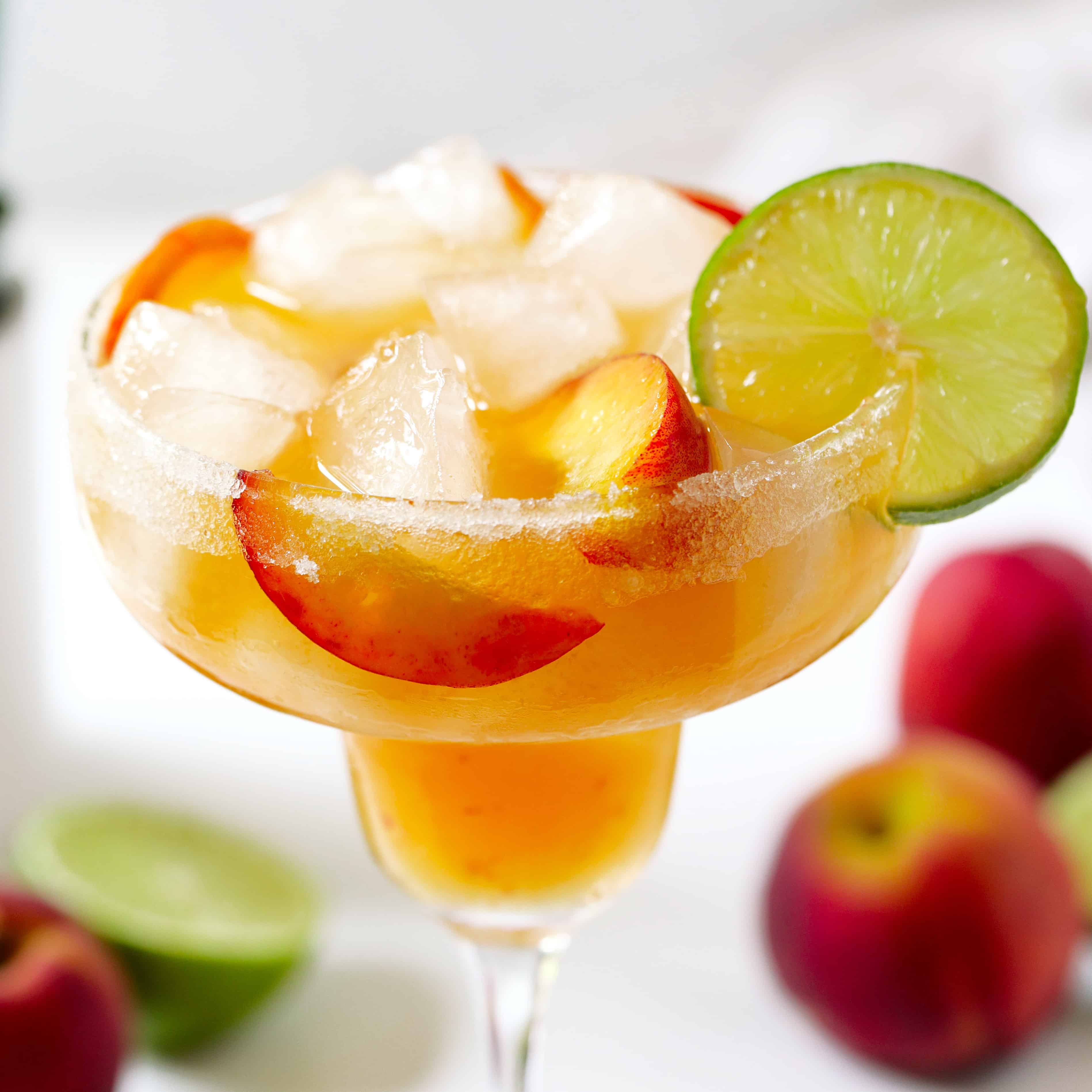 A peach margarita with the peach slices mixed in with the ice and a lime wheel around the edge of the glass. There are peaches and halved limes in the background.