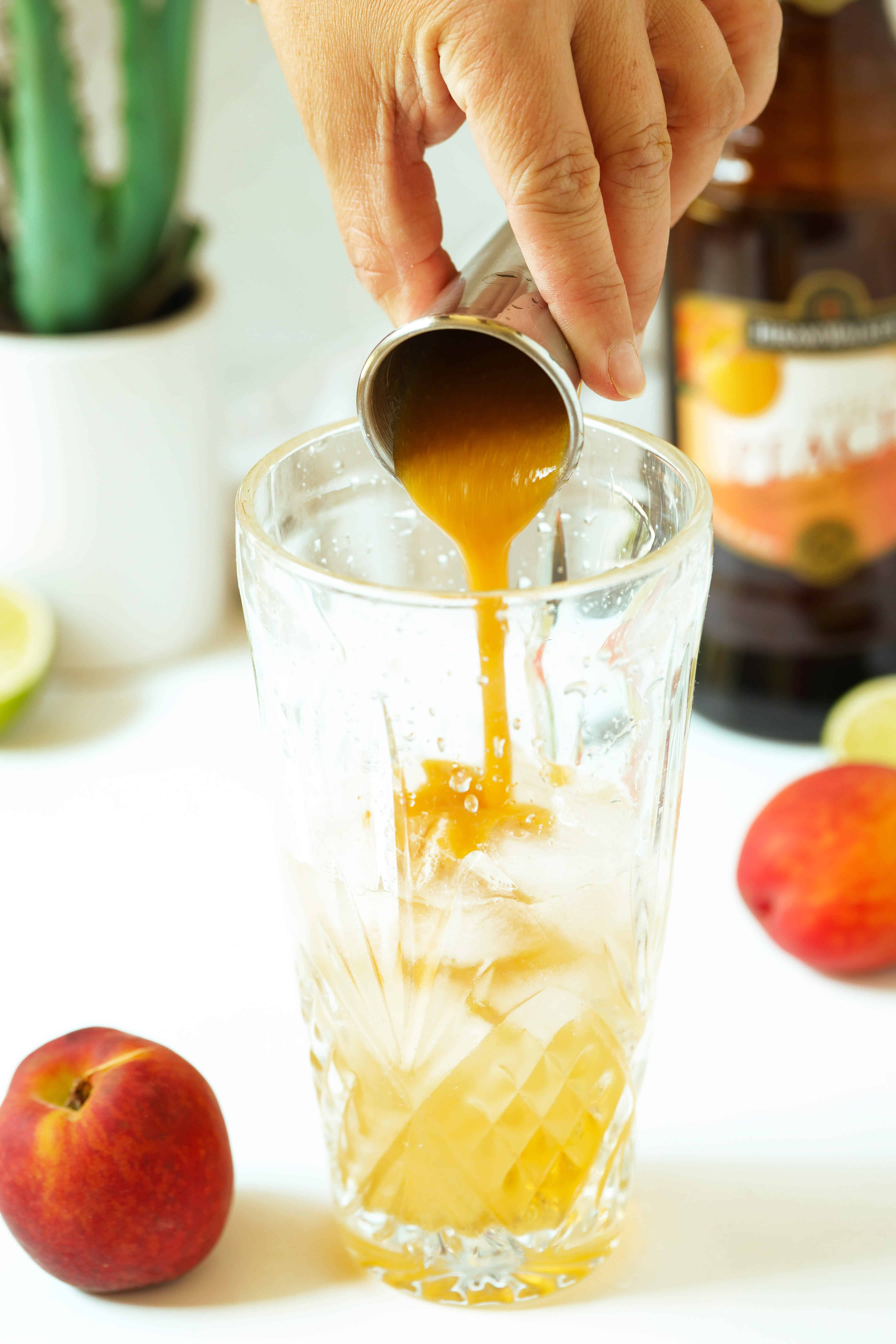 Peach purée being poured out of a jigger into a cocktail shaker. There is a bottle of peach schnapps, and agave plant, and peaches in the background.