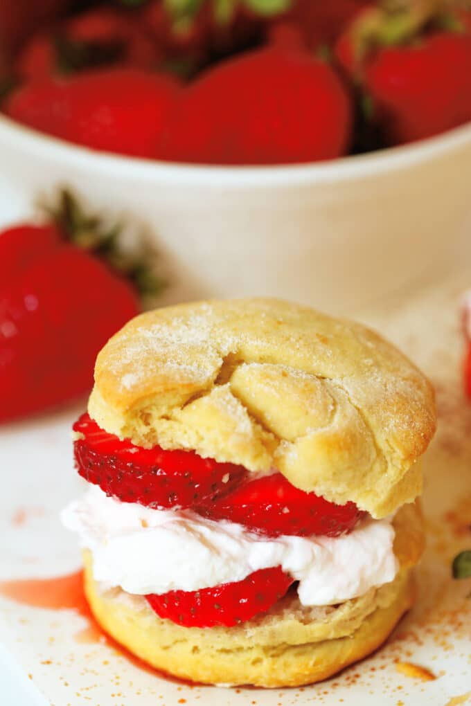 A fully assembled Strawberry Shortcake Biscuit with filling and whipped cream in the center and a bowl of fresh strawberries behind it.