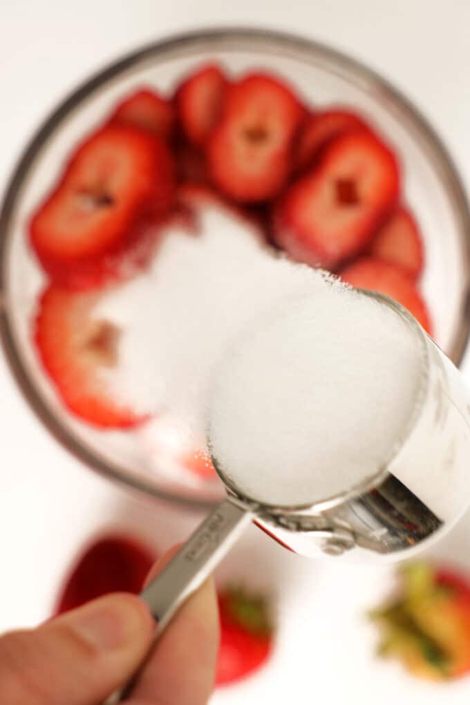 A measuring cup of sugar being poured into a bowl of sliced strawberries.