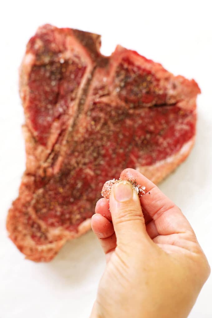 A hand sprinkling salt and pepper over a T-bone Steak on a white work surface.