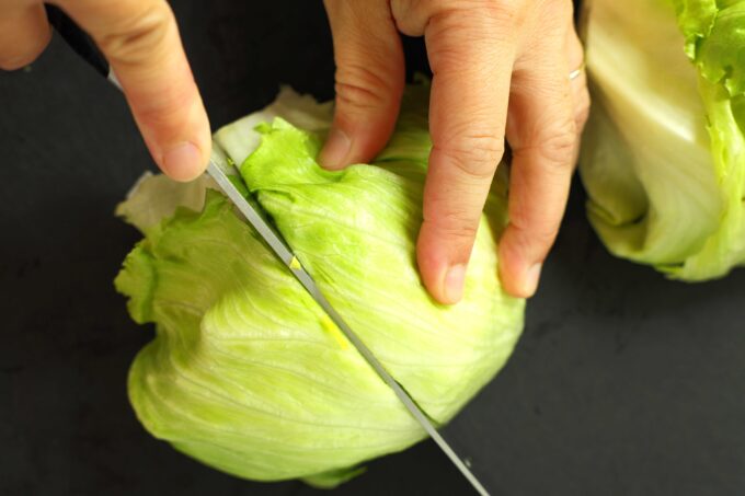 Hands using a large knife to cut a head of iceberg lettuce in half on a black cutting board.