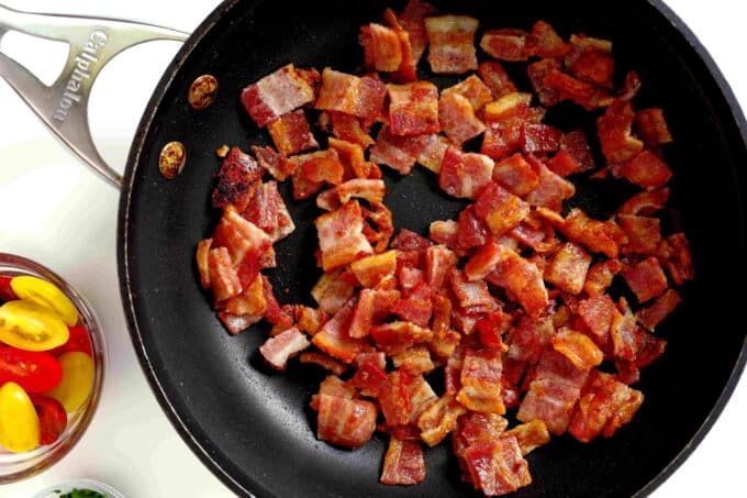 Little pieces of bacon cooking in a skillet with a small bowl of grape tomatoes nearby.