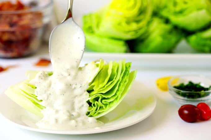 Blue cheese dressing being spooned onto a wedge of iceberg lettuce with more lettuce wedges and a bowl of cooked bacon pieces in the background.