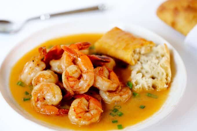 BBQ Shrimp on a white plate with a piece of baguette on the side. The pile of shrimp is surrounded by the golden sauce.