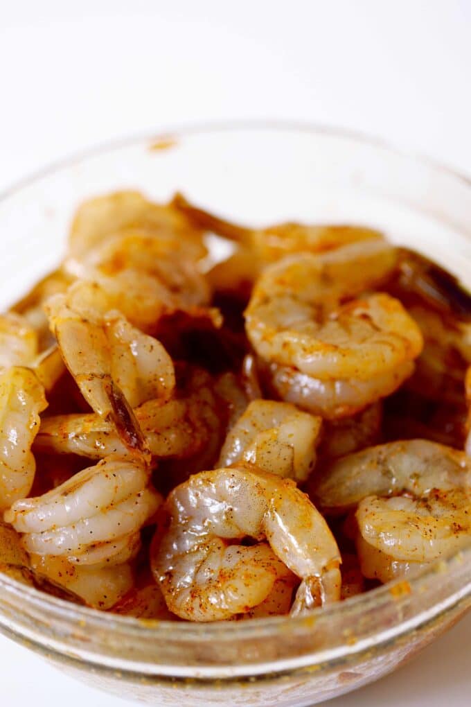 A glass mixing bowl full of large, raw shrimp that are coated in oil and seasoning.