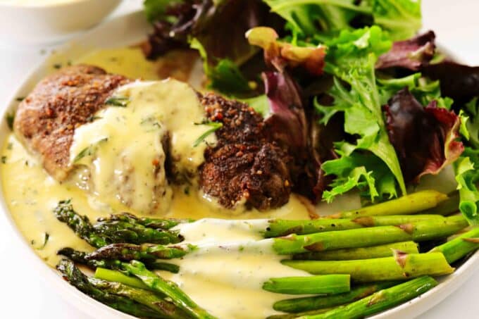A white dinner plate with Bearnaise Sauce poured over the steak and asparagus, and a salad on the side.