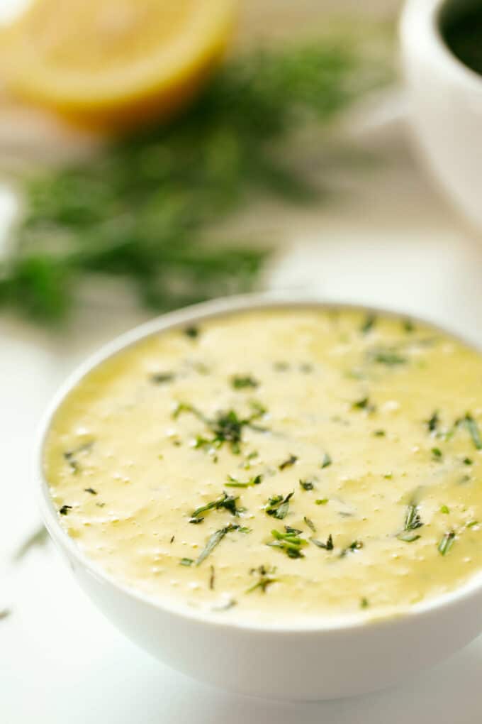 A bowl of Bearnaise Sauce sits on a white surface with fresh tarragon and half of a lemon in the background. The sauce is a pale yellow color with flecks of green. 