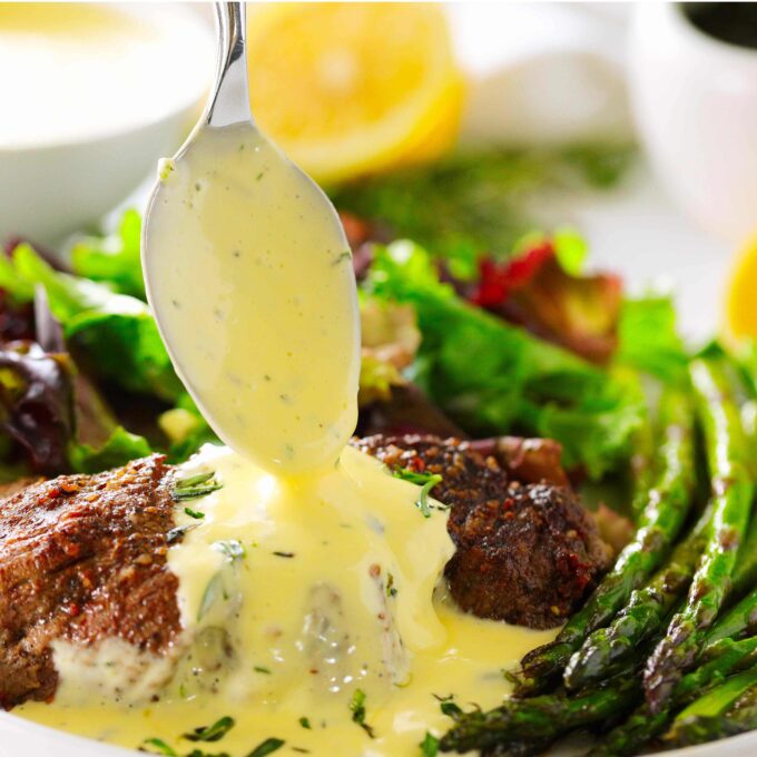 Bearnaise Sauce being spooned over a steak on a plate with a salad and asparagus.