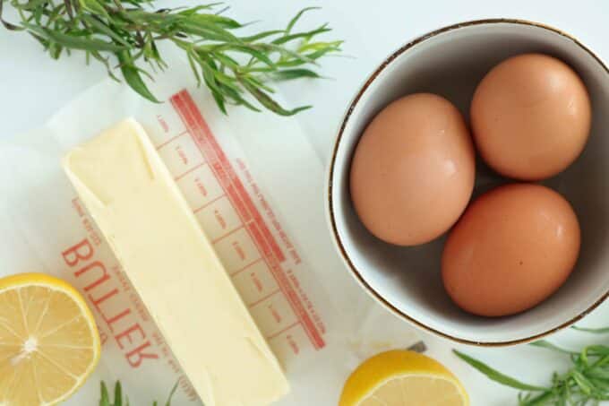 An overhead view of eggs, a stick of butter, a halved lemon, and fresh tarragon.