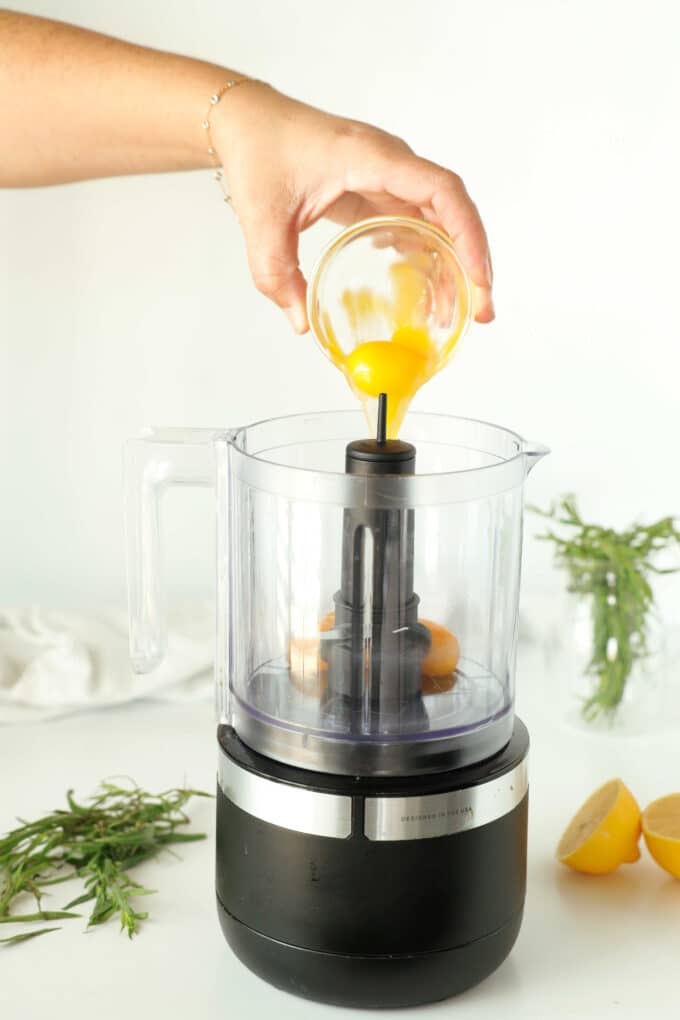 Egg yolks being dropped from a small bowl into a food processor.