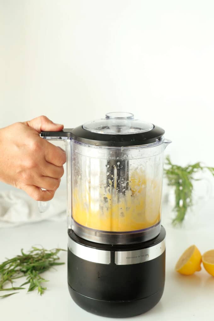 A hand holding down the button on a food processor as the water and egg yolks thicken inside.