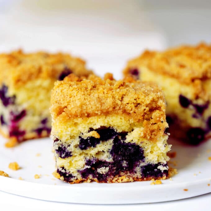 Several squares of Blueberry Coffee Cake sitting on a white plate. The cake is yellow with bursts of blueberries throughout and a golden topping.