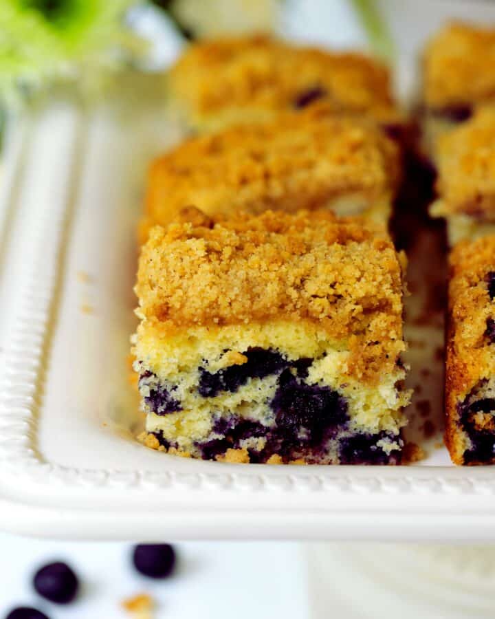 Several squares of Blueberry Coffee Cake sitting on a white cake stand. The cake is yellow with bursts of blueberries throughout and a golden topping.
