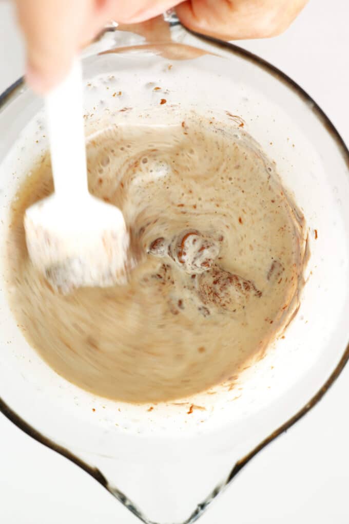 Melty chocolate chips being stirred into a measuring pitcher of heavy cream with a silicon spatula.