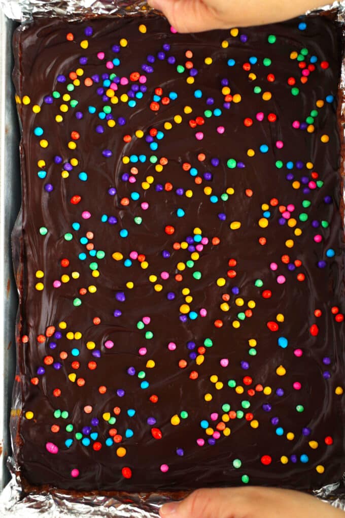 Hands using the edges of a foil lining to lift a pan of Cosmic Brownies out of the baking dish.