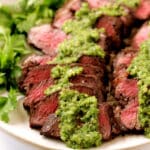 Two cooked Coulottle Steaks sliced up on a plate with parsley on the side and a green sauce spooned over the meat.