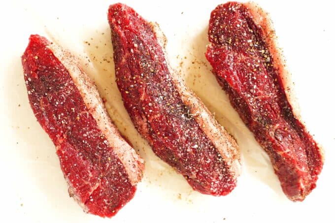 Three Coulotte Steaks that have been seasoned with salt and pepper, laying on a white surface.