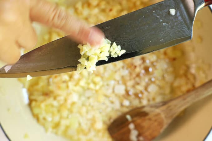 A hand pushing minced garlic off the side of a knife and into the pan of sautéed onion. 