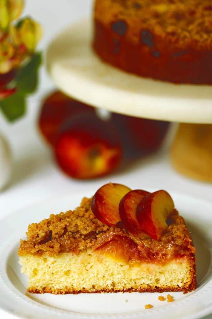 A piece of Peach Cake sitting on a white plate. It is a pale yellow color with a layer of peaches and a crumb topping on top. It is garnished with fresh peach slices. The rest of the cake on the cake stand and some whole peaches are sitting nearby.