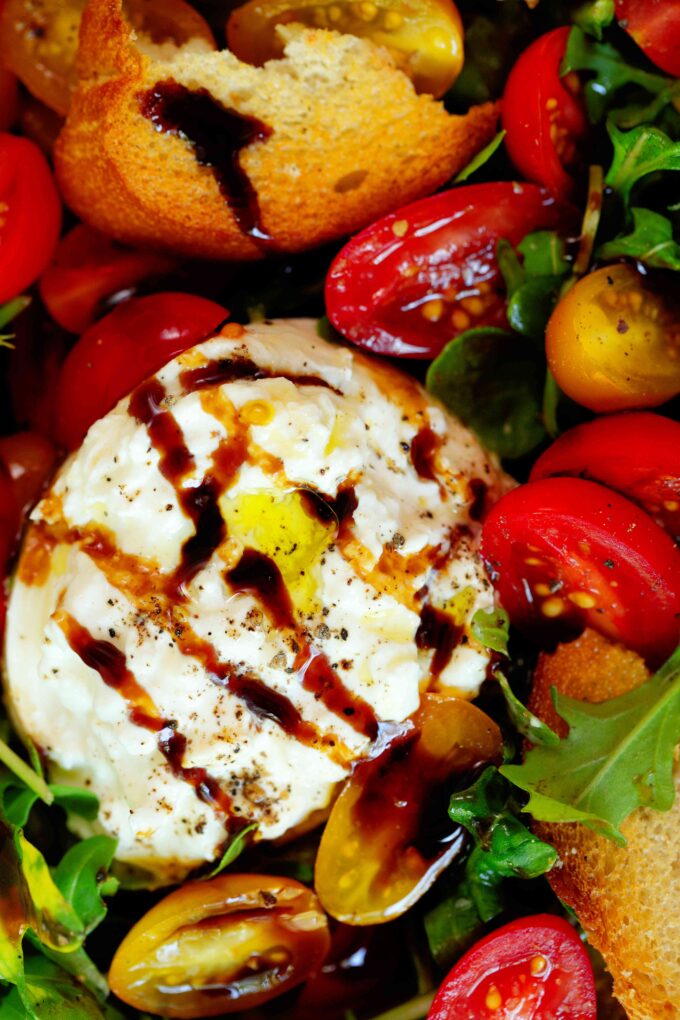 A close up, overhead look at a Burrata Salad. There is arugula, red and yellow tomatoes, burrata, and toasted baguette slices all topped off with olive oil and balsamic glaze.