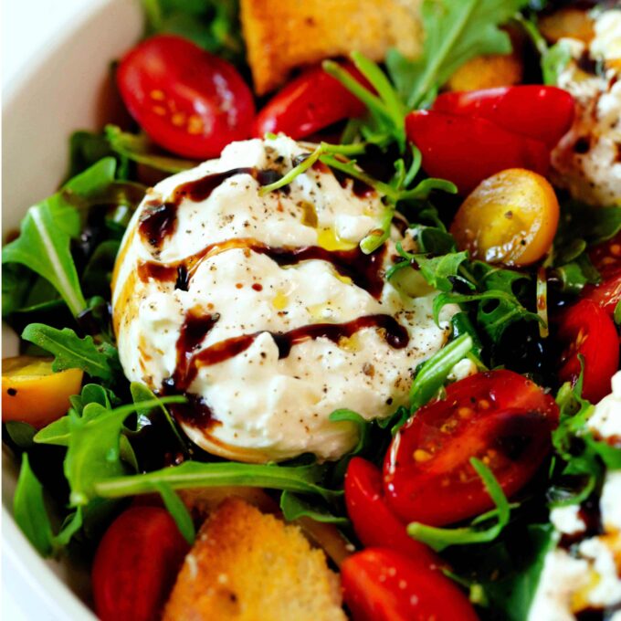 A close up of a white bowl of Burrata Salad. There is arugula, red and yellow tomatoes, burrata, and toasted baguette slices all topped off with olive oil and balsamic glaze.