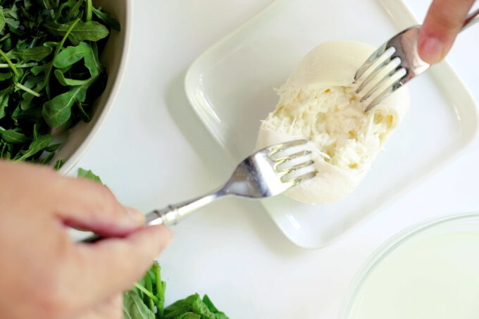 Hands using two forks to full a ball of burrata in half on a small, white plate.