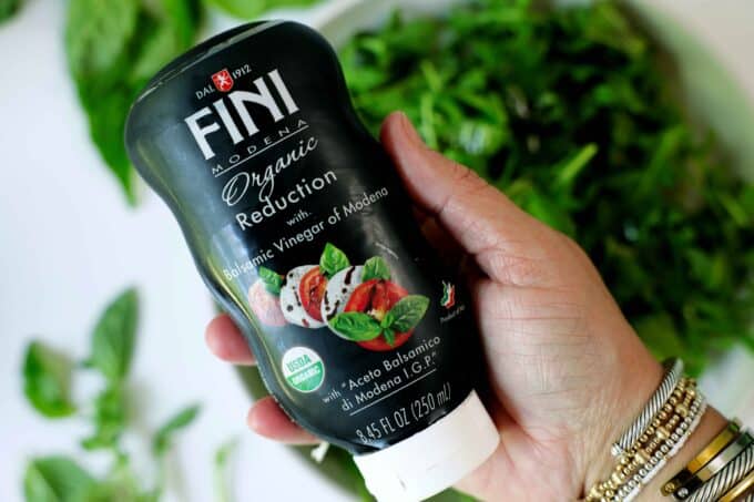 A hand holding a bottle of Fini brand balsamic reduction up to the camera, over a bowl of arugula.