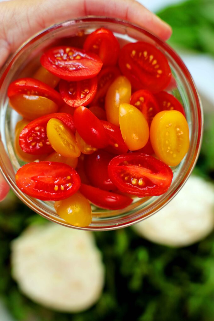 A hand holding a bowl of halved, yellow and red grape tomatoes up to the camera.