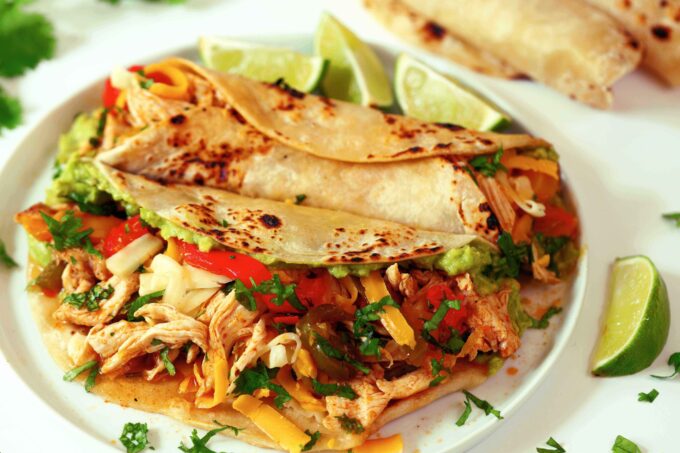 Two Crockpot Chicken Fajitas on a white plate with lime wedges on the side and cilantro sprinkled over top.