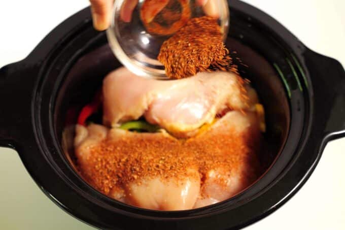 A hand shaking a spice mix from a small bowl onto raw chicken breasts in a crockpot.