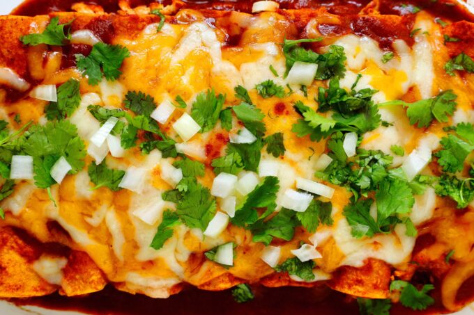 An overhead view of a pan of enchiladas topped with red Enchilada Sauce, melty cheese, chopped onion, and cilantro.