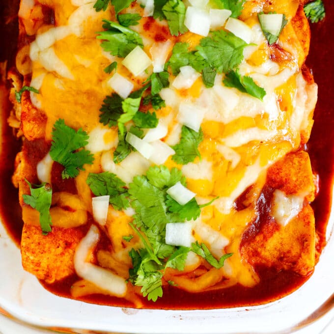 A close up, overhead view of a pan of enchiladas topped with red Enchilada Sauce, melty cheese, chopped onion, and cilantro.