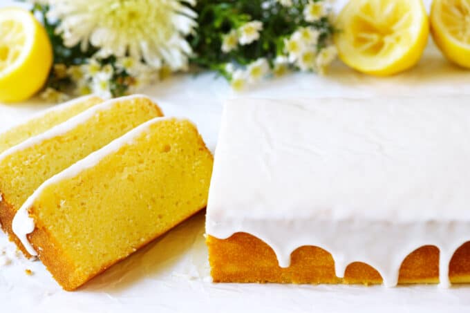 A Lemon Pound Cake covered in a thick, white glaze, with a couple of pieces sliced off of one end and some flowers and halved lemons sitting behind it.